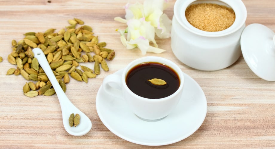 Café com cardamomo servido em uma xícara branca em uma mesa de madeira clara. Ao lado, grãos de cardamomo estão espalhados.