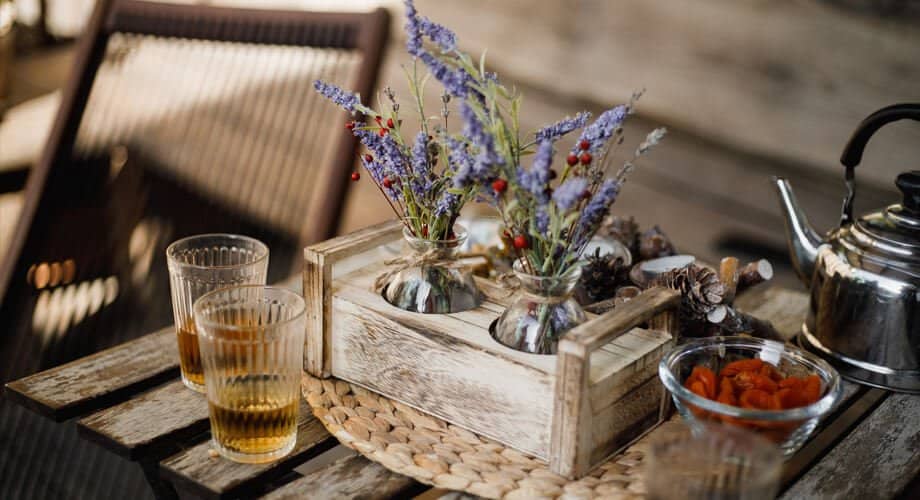 Cantinho do café simples com flores roxas em um suporte de madeira, sobre uma mesa de madeira. Ao lado, há dois copos com café.