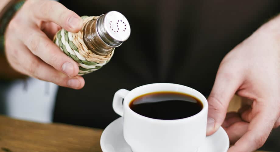 Café com sal sendo servido em uma xícara branca. Mão masculina segura recipiente de sal e o leva em direção à xícara.