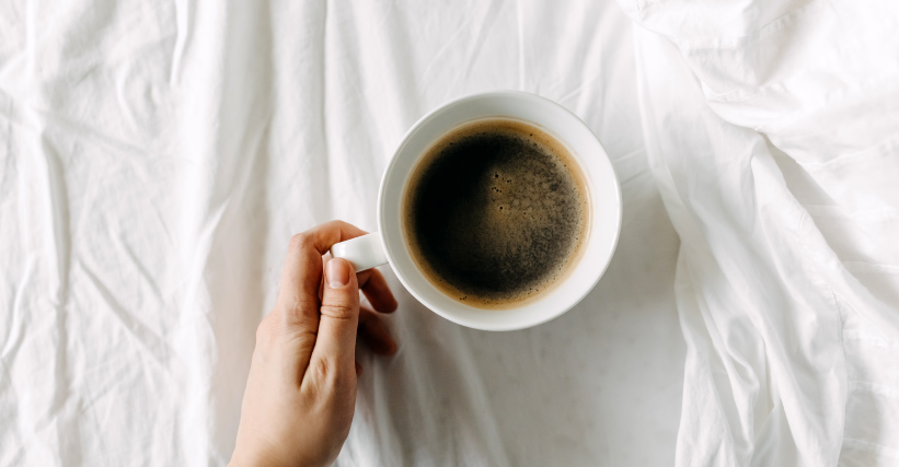 Café tira o sono? Entenda os efeitos da cafeína! Café tira o sono? Sobre lençóis brancos, vemos uma mão segurando uma xícara de café.