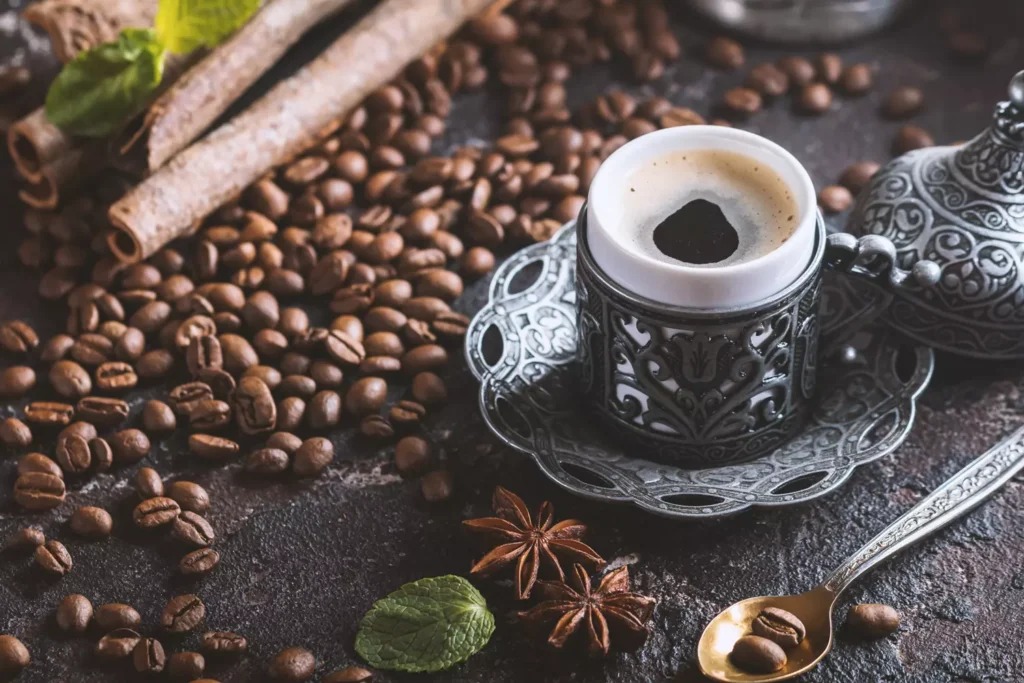 xícara de café sobre uma bandeja metálica, rodeada de grãos de café, paus de canela, folhas de hortelã e estrelas de anis